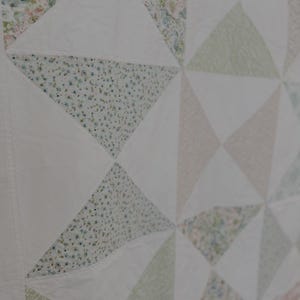 May include: Close-up of a handmade quilt with a geometric pattern. The quilt features white, floral, and solid triangles. The floral triangles have a light blue, pink, and green color palette. The quilt is made of soft fabric.