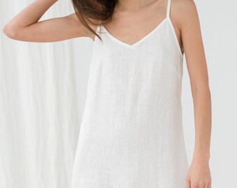 Puede incluir: Un vestido corto blanco sin mangas con escote en V y tirantes finos. La prenda está confeccionada con un tejido texturizado que cae en una silueta recta y relajada, terminando por encima de las rodillas con un dobladillo sencillo.