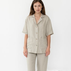 Pu&ograve; includere: Una persona indossa un pigiama in lino color beige chiaro con una camicia con colletto e pantaloni. La camicia ha una chiusura con bottoni sul davanti e una tasca a toppa sul lato sinistro. I pantaloni hanno una gamba dritta.