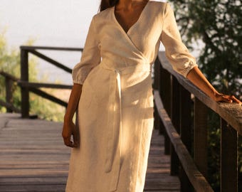 May include: A woman wearing a white linen wrap dress stands on a wooden walkway. The dress has a V-neckline and a tie belt. She is barefoot and has long brown hair.