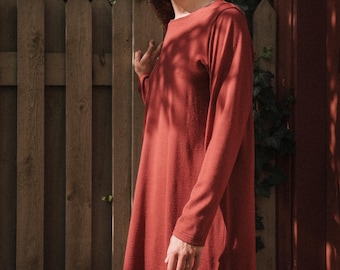 May include: A rust-colored long-sleeve dress with a round neckline. The dress is made of a soft, flowing fabric and falls to about mid-thigh. The model is standing in front of a wooden fence.