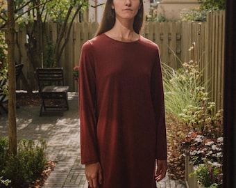 May include: A long-sleeved, burgundy-colored dress. The dress has a simple, round neckline and a relaxed fit. The person is standing in a garden setting with a wooden fence and trees in the background.
