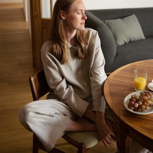 Può includere: Una donna con una camicia e pantaloni di lino beige chiaro è seduta su una sedia di legno a un tavolo con un bicchiere di succo d'arancia e una ciotola di uva.