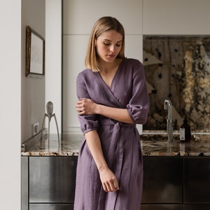 May include: A woman wearing a long, purple linen wrap dress with 3/4 sleeves. The dress has a tie belt at the waist. She is standing in a kitchen with a stainless steel countertop and cabinets.