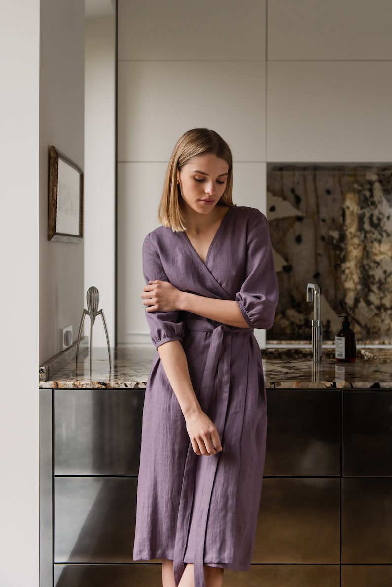 May include: A woman wearing a long, purple linen wrap dress with 3/4 sleeves. The dress has a tie belt at the waist. She is standing in a kitchen with a stainless steel countertop and cabinets.