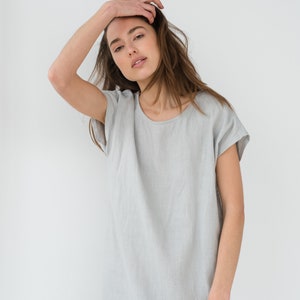 May include: A woman wearing a light gray linen dress with short sleeves. She is standing in front of a white background.