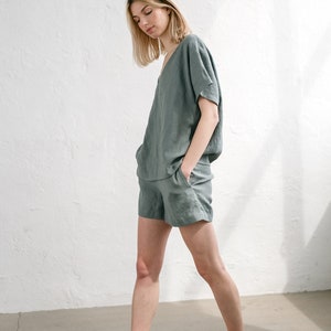 May include: A woman wearing a sage green linen short set. The set includes a v-neck top with short sleeves and matching shorts. The woman is standing on a concrete floor.