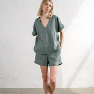 May include: A woman wearing a sage green linen top and matching shorts. The top has a V-neck and short sleeves. The shorts are a relaxed fit with a wide leg.