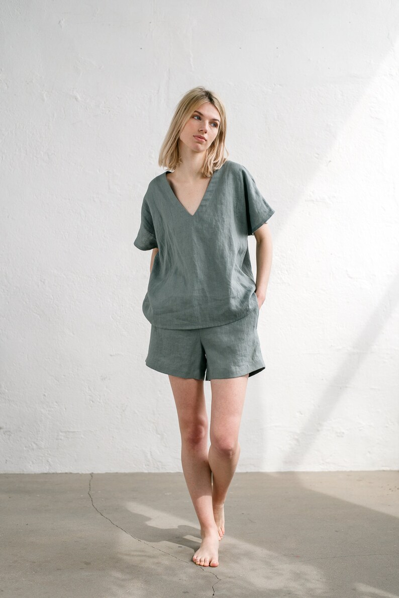 May include: A woman wearing a sage green linen top and matching shorts. The top has a V-neck and short sleeves. The shorts are a relaxed fit with a wide leg.