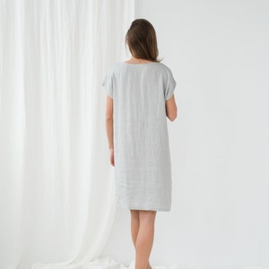 May include: A woman wearing a light gray linen dress with short sleeves. She is standing on a white surface with a white curtain in the background.