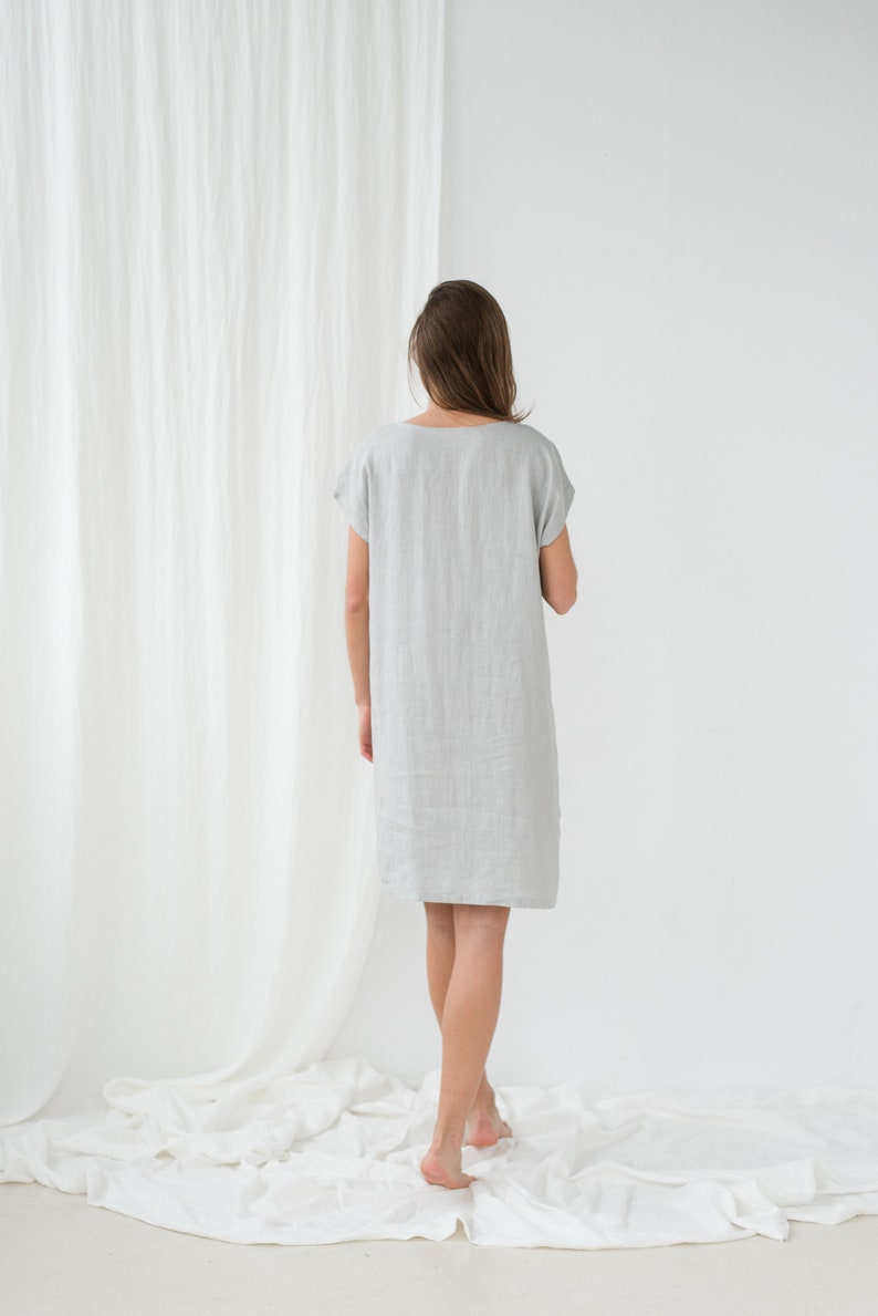 May include: A woman wearing a light gray linen dress with short sleeves. She is standing on a white surface with a white curtain in the background.