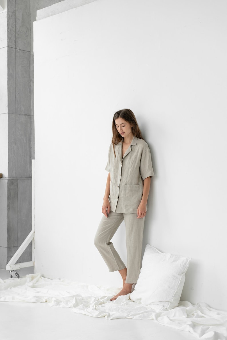Pu&ograve; includere: Una donna indossa un pigiama in lino beige chiaro con una camicia con colletto e pantaloni. &Egrave; in piedi in una stanza bianca con un cuscino bianco sul pavimento.