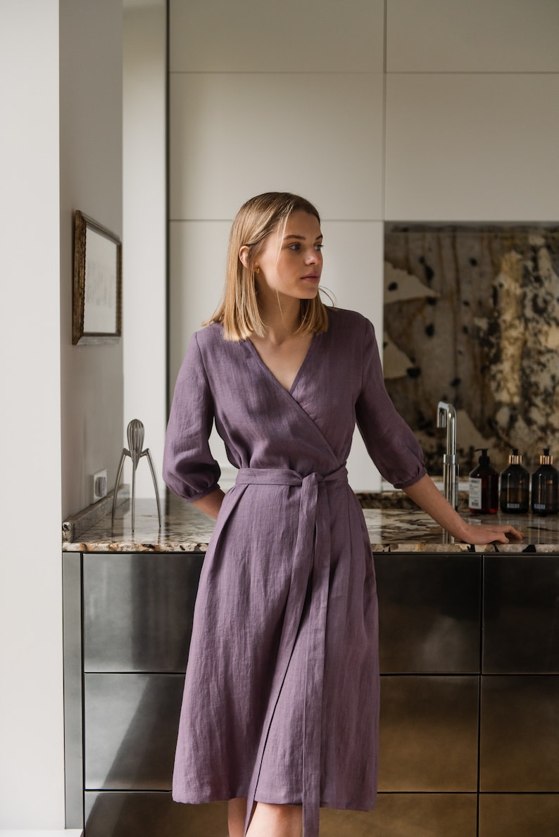 May include: A woman wearing a purple linen wrap dress with a self-tie belt. The dress has a V-neckline and 3/4 sleeves.