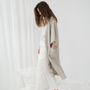 Peut inclure: Une femme portant une robe en lin gris clair sur un haut et un pantalon en lin blanc. Elle se tient debout sur un drap blanc avec un oreiller blanc derri&egrave;re elle.