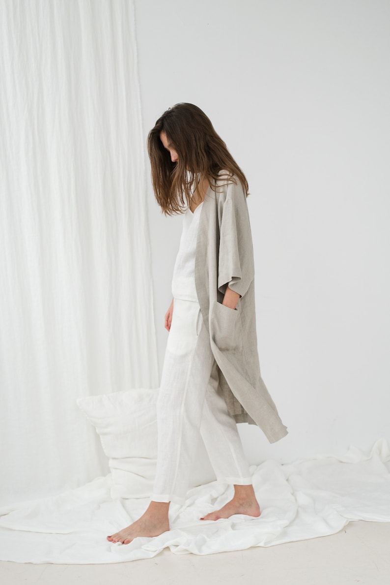 Peut inclure: Une femme portant une robe en lin gris clair sur un haut et un pantalon en lin blanc. Elle se tient debout sur un drap blanc avec un oreiller blanc derri&egrave;re elle.