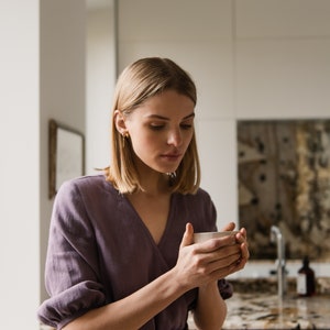 May include: A woman in a purple linen dress holds a white ceramic mug in her hands. She is looking down at the mug and smiling.