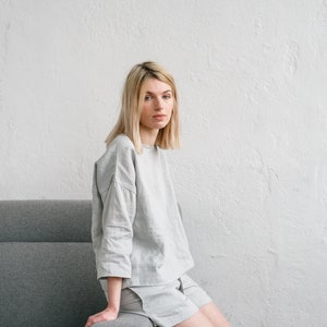 May include: A person wearing a light grey linen top and shorts sits on a grey sofa. The person has blonde hair and is looking at the camera.