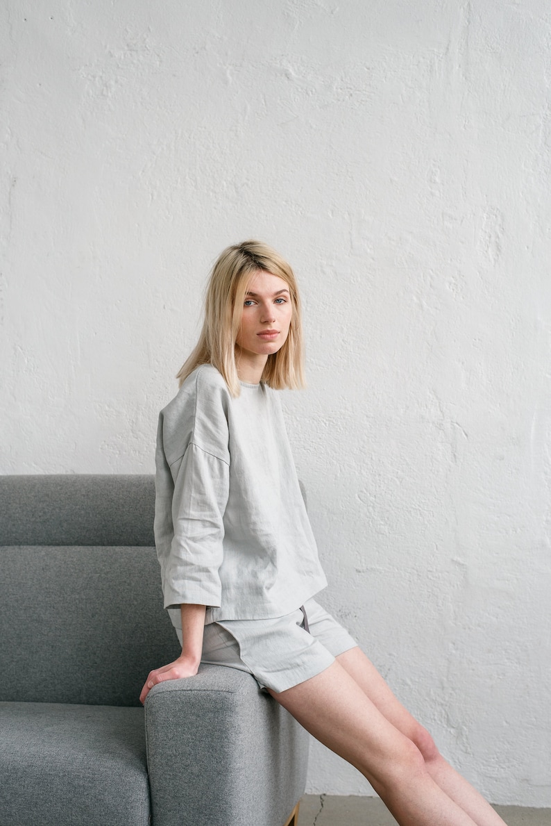 May include: A person wearing a light grey linen top and shorts sits on a grey sofa. The person has blonde hair and is looking at the camera.