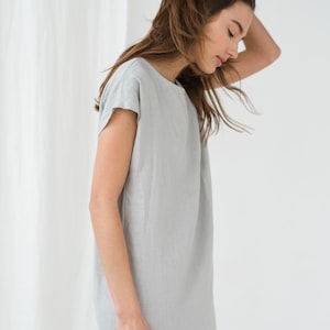 May include: A woman wearing a light gray linen dress with short sleeves. The dress is simple and loose-fitting, with a scoop neckline.