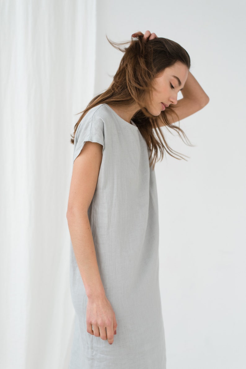 May include: A woman wearing a light gray linen dress with short sleeves. The dress is simple and loose-fitting, with a scoop neckline.