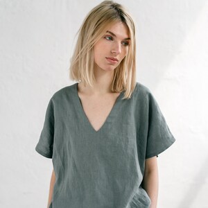 May include: A woman wearing a loose-fitting, sage green linen top with a v-neck and short sleeves. The top is made of a soft, breathable fabric and has a relaxed fit.