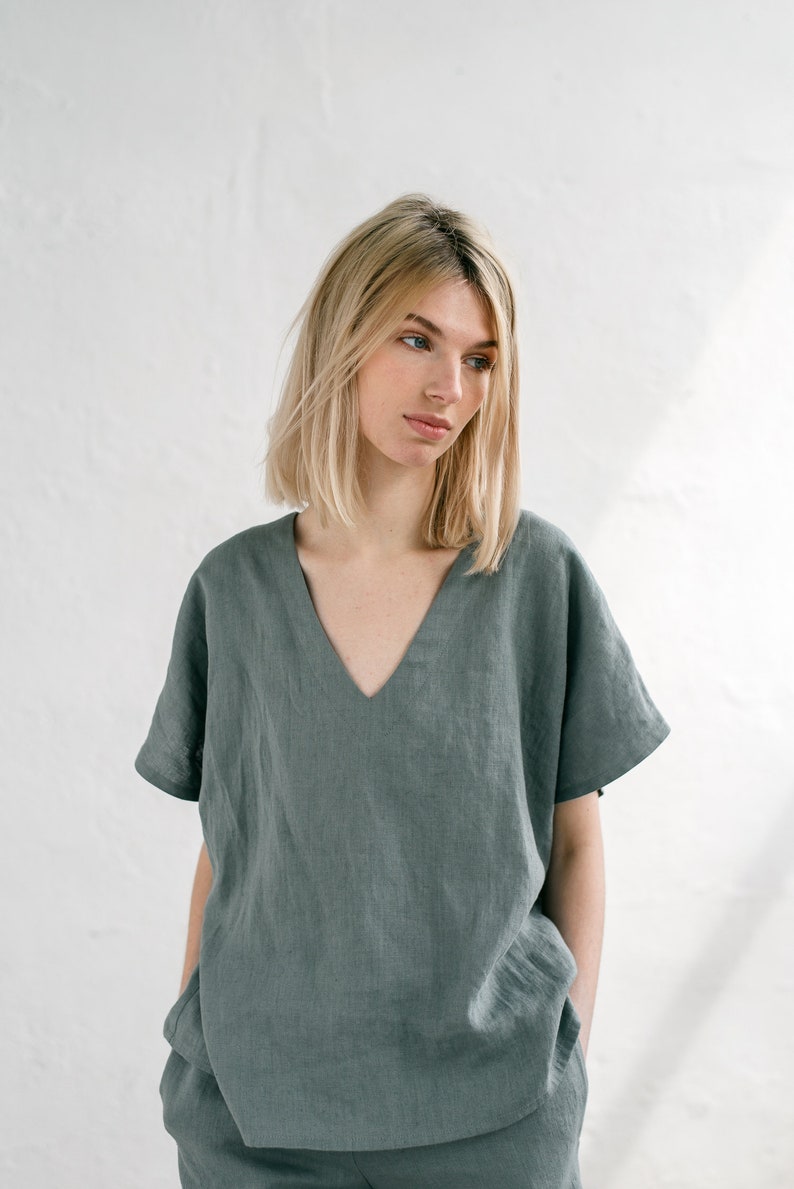 May include: A woman wearing a loose-fitting, sage green linen top with a v-neck and short sleeves. The top is made of a soft, breathable fabric and has a relaxed fit.