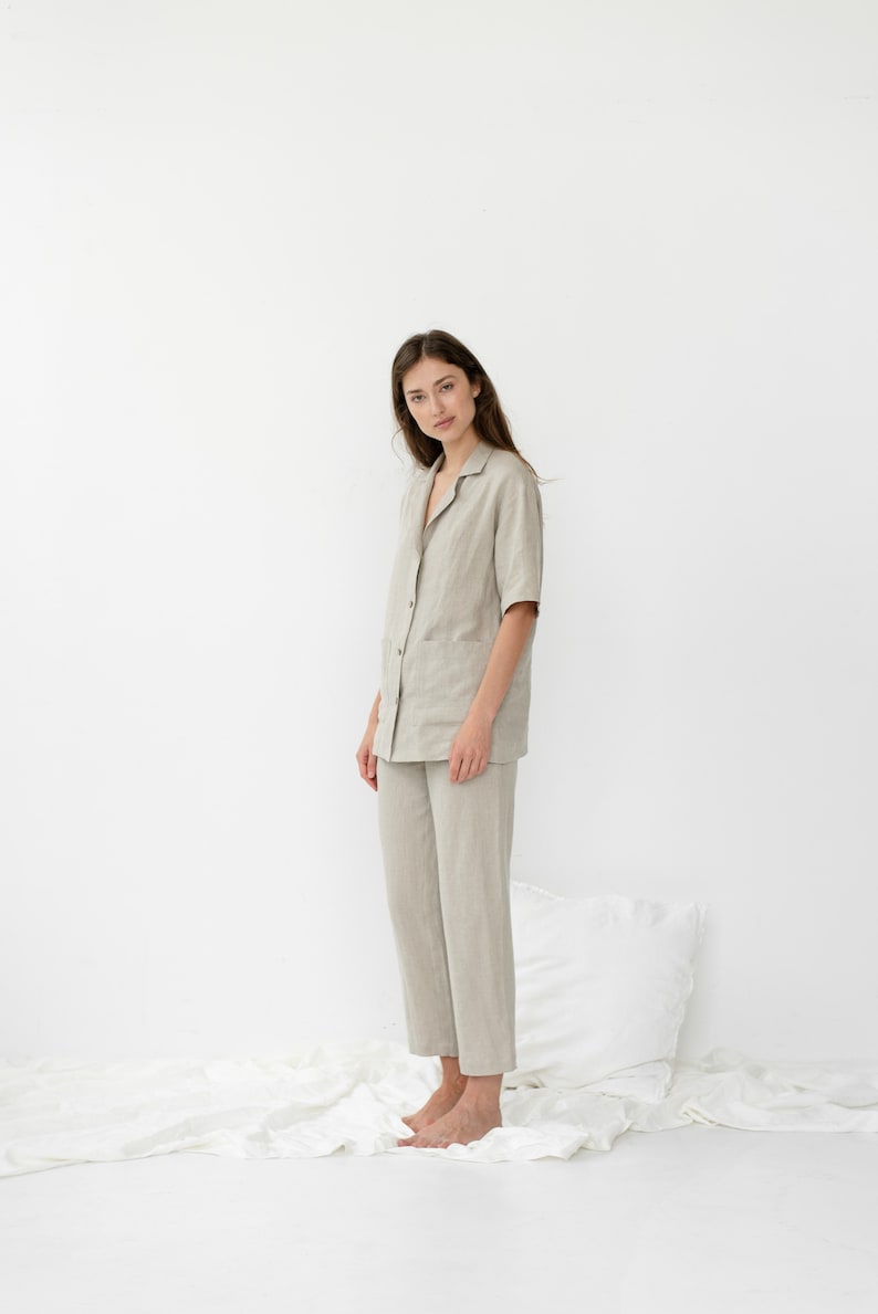 Pu&ograve; includere: Una persona indossa un pigiama in lino beige chiaro con una camicia con colletto e pantaloni. La persona &egrave; in piedi su un lenzuolo bianco.