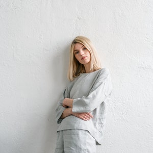 May include: A woman wearing a light grey linen top and shorts set. The top has a relaxed fit and the shorts are a bit shorter. She is standing against a white wall.