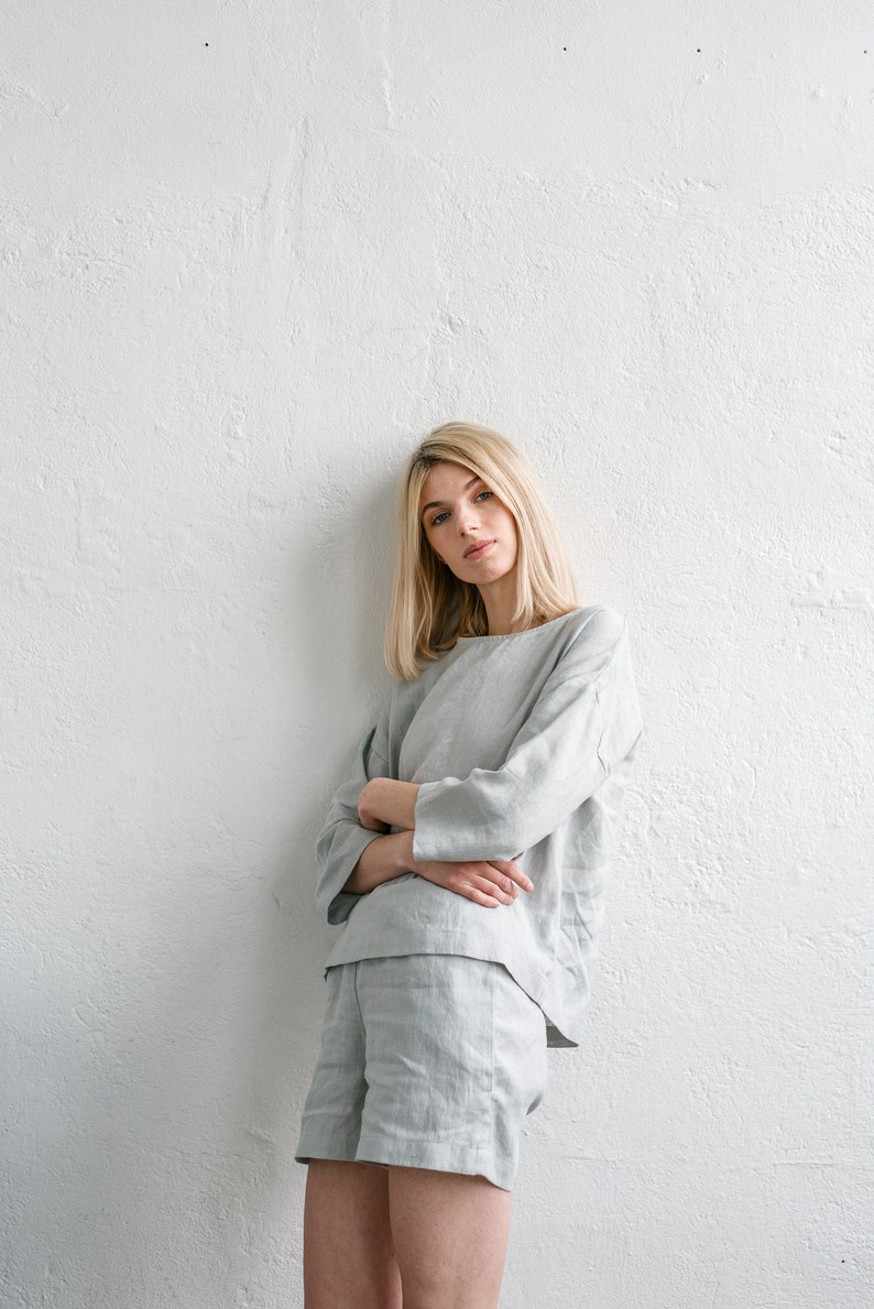 May include: A woman wearing a light grey linen top and shorts set. The top has a relaxed fit and the shorts are a bit shorter. She is standing against a white wall.