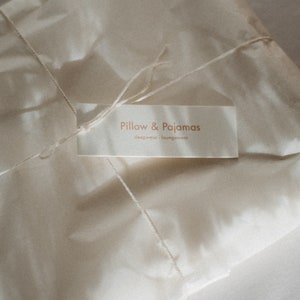 Puede incluir: Un paquete de papel blanco atado con una cuerda y una etiqueta que dice "Pillow & Pajamas, sleepwear + loungewear".