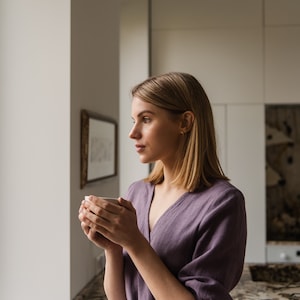May include: A woman in a purple linen dress holds a white mug and looks out a window.