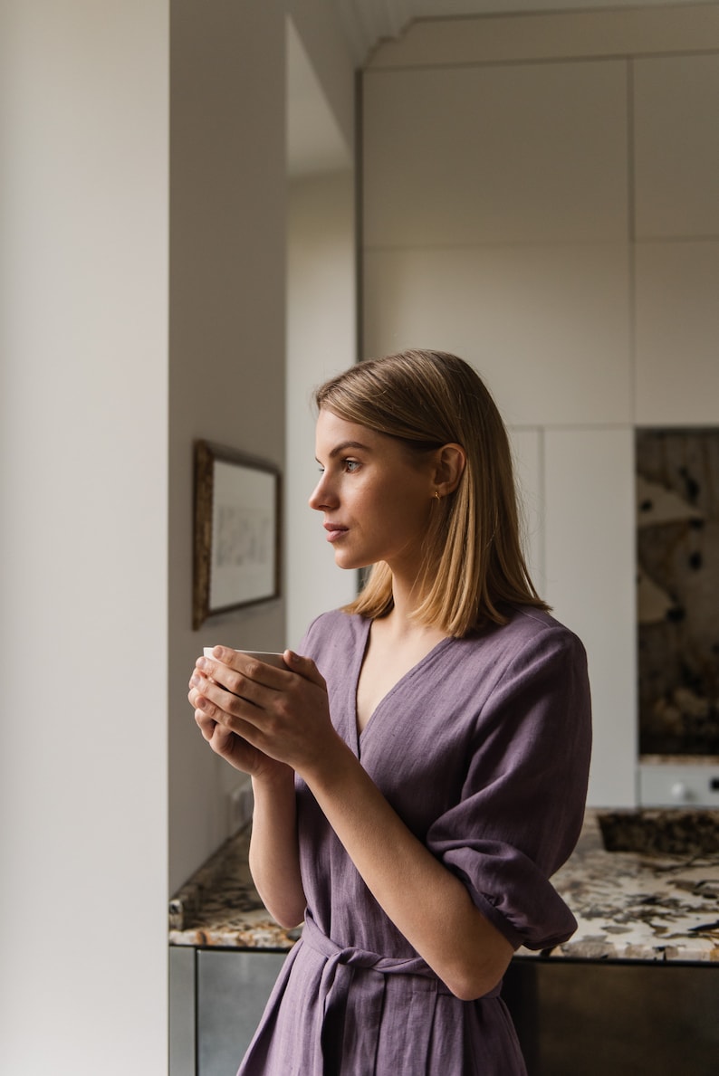 May include: A woman in a purple linen dress holds a white mug and looks out a window.