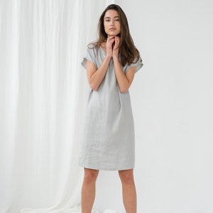 May include: A woman wearing a light gray linen dress with short sleeves. She is standing on a white sheet with a white curtain in the background.