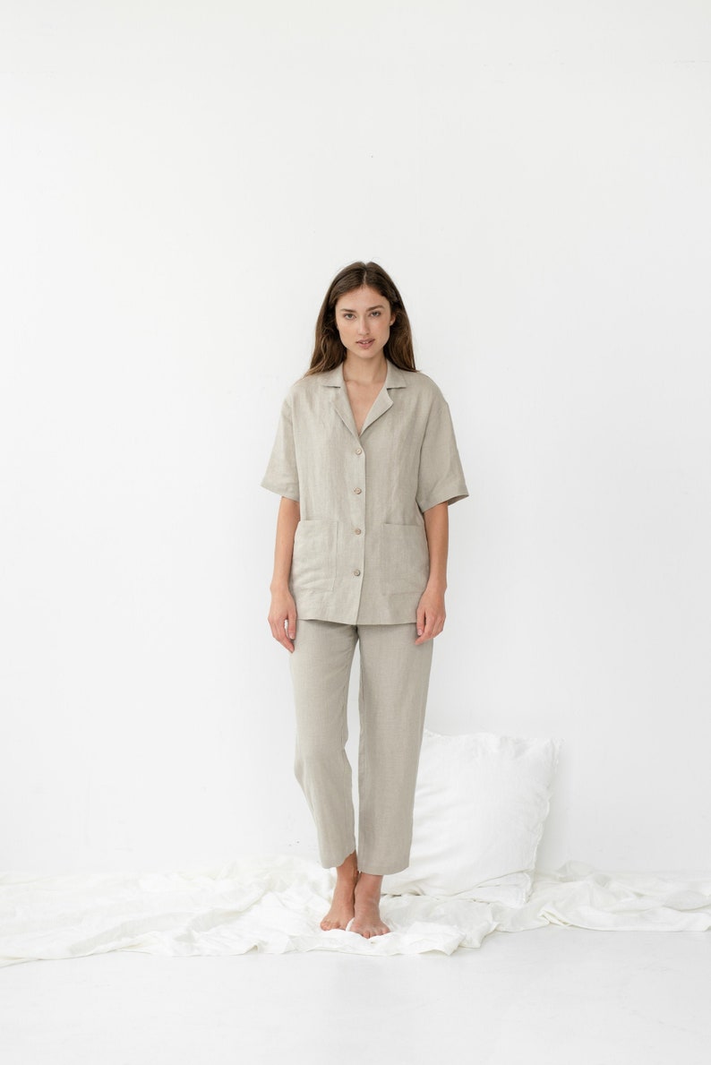 Pu&ograve; includere: Una persona indossa un pigiama in lino color beige chiaro con una camicia con colletto e pantaloni. La camicia ha una chiusura con bottoni sul davanti e una tasca a toppa sul lato sinistro. I pantaloni hanno una gamba dritta.