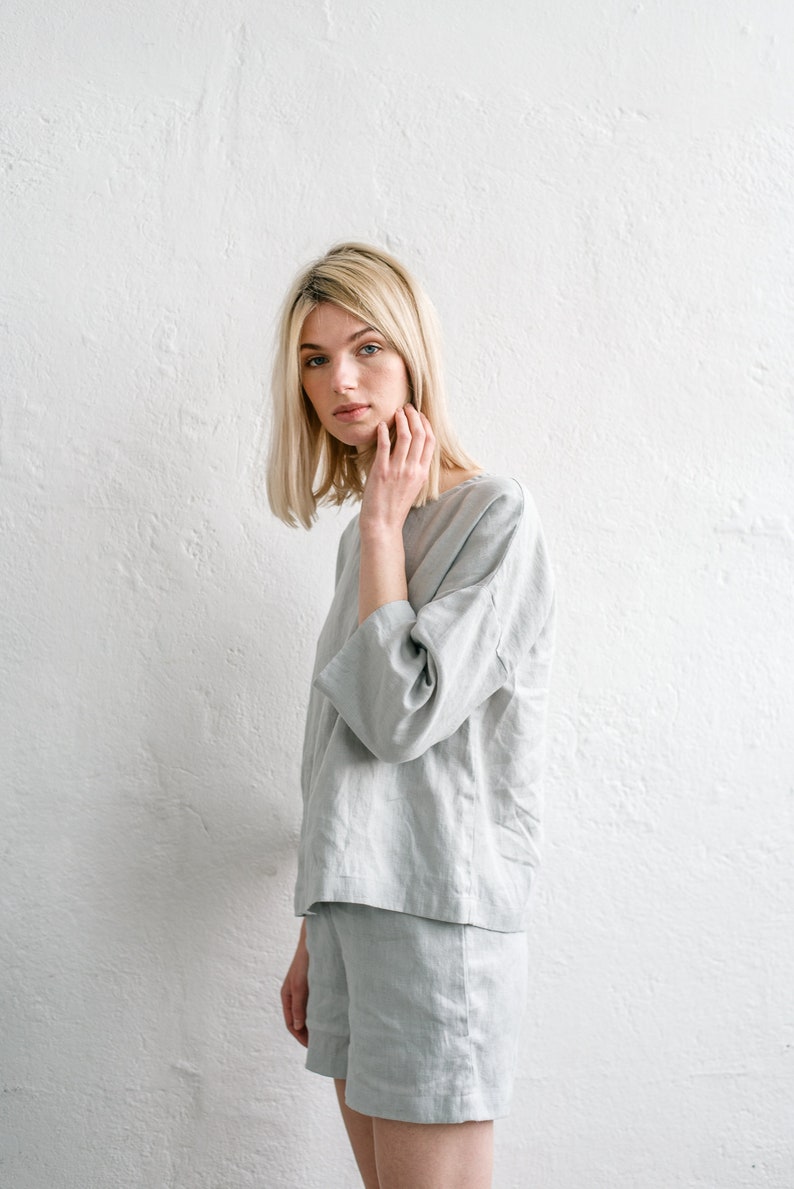 May include: A woman wearing a light grey linen top and matching shorts. The top has a relaxed fit and three-quarter sleeves. The shorts are a bit shorter and have a straight leg.