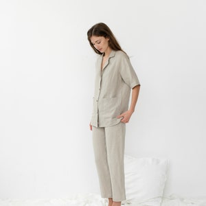 Pu&ograve; includere: Una persona indossa un pigiama in lino beige chiaro con una camicia con colletto e pantaloni. La persona &egrave; in piedi su un lenzuolo bianco.