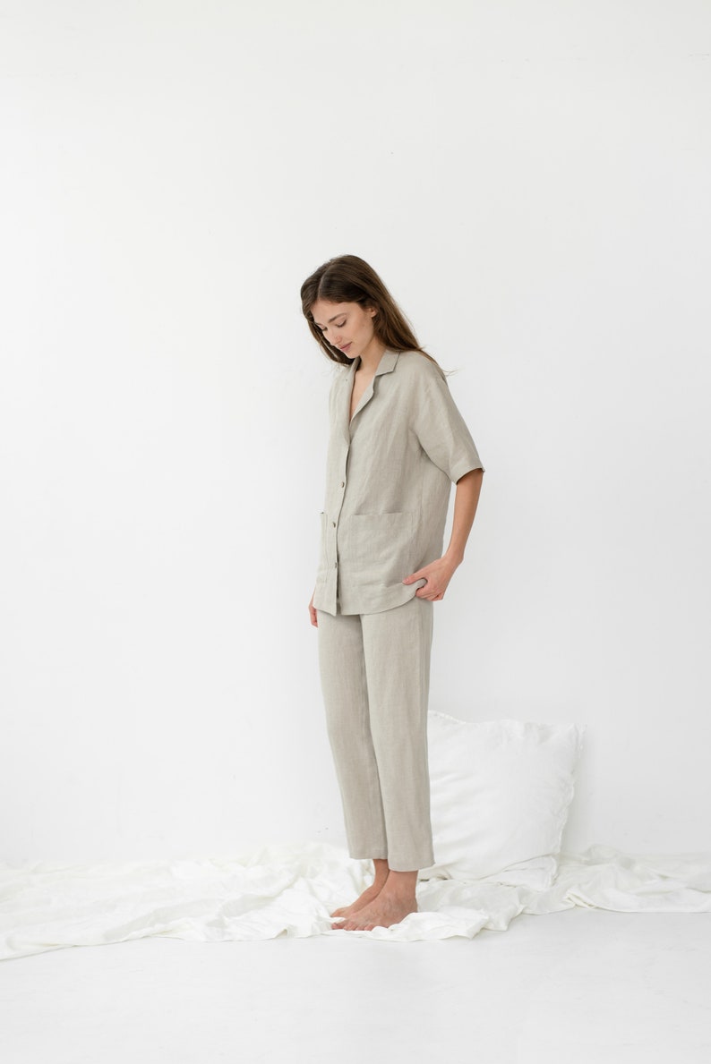 Pu&ograve; includere: Una persona indossa un pigiama in lino beige chiaro con una camicia con colletto e pantaloni. La persona &egrave; in piedi su un lenzuolo bianco.