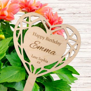 May include: A wooden heart-shaped plant stake, engraved with "Happy Birthday Emma" and "Love Sophia", featuring an openwork design. The stake is set amongst green foliage and vibrant pink flowers, creating a decorative garden accent.