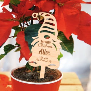 Op de afbeelding: Een houten kerstkabouter plantenstaak met de tekst "Merry Christmas Alice". De staak staat in een rode pot met een poinsettia plant. De woorden "Lots of love Lily, David and Ivy" zijn ook gegraveerd.