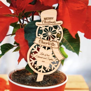 Könnte beinhalten: Ein Weihnachts-Pflanzschild aus Holz in Form eines Schneemanns. Es hat einen Hut, ein Schneeflocken-Design und einen Schal mit dem Namen "Charlotte". Der Text "Merry Christmas" und "Lots of love Edward and Sophia" sind ebenfalls sichtbar. Der Pflanzschild befindet sich in einem roten Topf mit einer Poinsettie.
