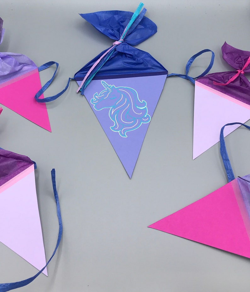 Pu&ograve; includere: Un set di bandierine triangolari per feste di colore viola, rosa e bianco con un design di unicorno su una delle bandierine. Le bandierine sono legate insieme da un nastro blu.