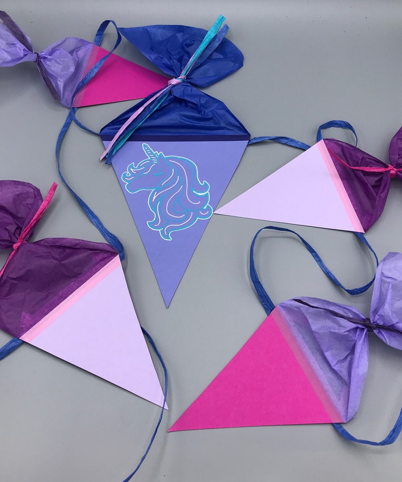 Pu&ograve; includere: Uno striscione triangolare viola, rosa e blu con una silhouette di unicorno su uno dei triangoli. Lo striscione &egrave; fatto di carta velina ed &egrave; infilato con un nastro blu.