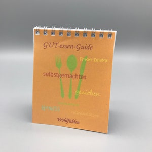 Könnte beinhalten: Ein kleines orangefarbenes Heft mit Spiralbindung. Der Umschlag trägt den Text "GUT-essen-Guide" und zeigt eine Gabel, ein Messer und einen Löffel mit grünen Blättern. Der Text "Frische Zutaten", "selbstgemachtes", "genießen", "Genuss", "Geschmackvoll" und "Wohlfühlen" befindet sich ebenfalls auf dem Umschlag.