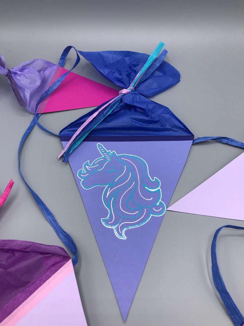 Pu&ograve; includere: Uno striscione triangolare viola e rosa con un'illustrazione di unicorno blu. Lo striscione &egrave; decorato con nastri blu, rosa e turchese.