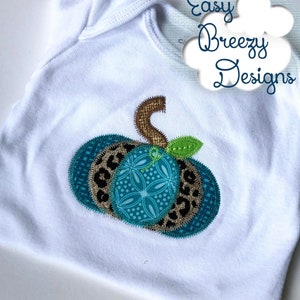 Könnte beinhalten: Ein weißer Baby-Strampler mit einer Applikation aus einer türkisfarbenen, braunen und schwarzen Patchwork-Kürbis. Der Text "Easy Breezy Designs" ist in der oberen rechten Ecke des Bildes sichtbar.