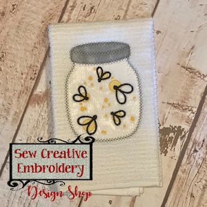 May include: White kitchen towel with an embroidered design of a glass jar filled with fireflies. The jar has a gray lid and the fireflies are yellow with black outlines. The text "Sew Creative Embroidery Design Shop" is at the bottom of the towel.