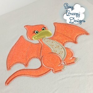 May include: Embroidered design of a pink and orange dinosaur with a yellow beak and green eyes. The dinosaur has a glittery finish and is flying with its wings spread out.