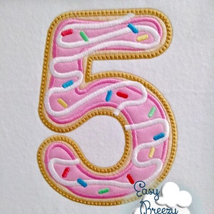 Könnte beinhalten: Rosaer gestickter Patch in Donutform mit weißem Zuckerguss und bunten Streuseln. Die Zahl 5 ist mit weißem Zuckerguss umrandet. Der Donut ist in Gold umrandet.