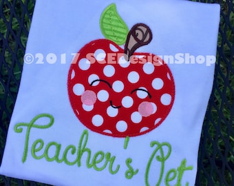 May include: White t-shirt with a red and white polka dot apple with a green leaf and brown stem. The apple has a smiling face with pink cheeks. The text "Teacher's Pet" is embroidered in green below the apple.