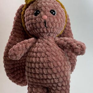 May include: A crocheted plush bunny toy with a pink body and a yellow headband. The bunny has black eyes and a pink nose.